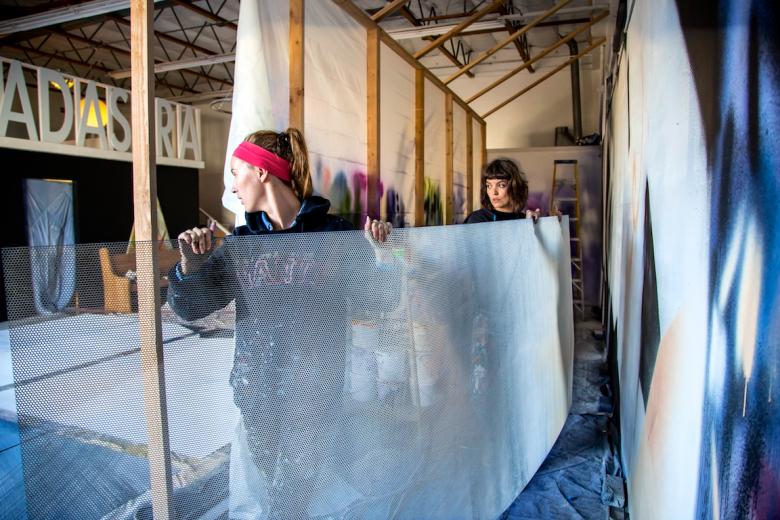 Hennessy Christophel (left) and Sofia Lacin (right) carry a panel of perforated metal from their studio. The two muralists created work for the WAL courtyard and alley Hennessy Christophel (left) and Sofia Lacin (right) carry a panel of perforated metal from their studio. The two muralists created work for the WAL courtyard and alley