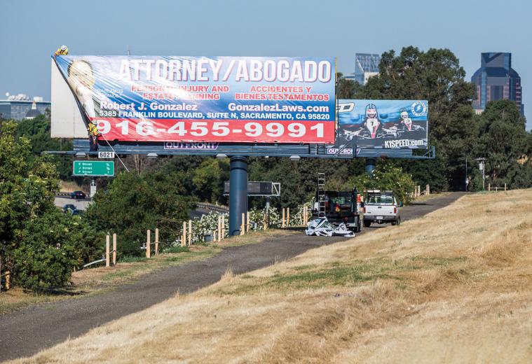 On this morning, the men first pull down an old vinyl (billboard speak for “sign”) before installing the new one — advertising a local law firm — as commuters speed past on the road below.  On this morning, the men first pull down an old vinyl (billboard speak for “sign”) before installing the new one — advertising a local law firm — as commuters speed past on the road below.